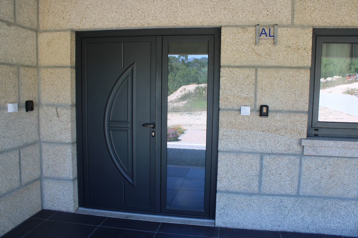 A modern entrance features a sleek black door with a unique curved design. The door is framed by textured stone walls, and a window adjacent to it allows natural light to enter. A paved walkway is visible in the background, leading to the surrounding landscape.