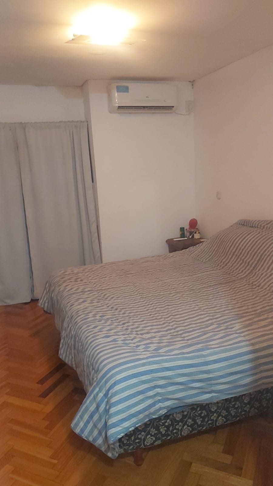 A spacious sleeping area features a striped blanket draped over a large bed. Light wood flooring complements the room, while a small nightstand holds personal items. A window is partially covered by light curtains, and an air conditioning unit is mounted on the wall.