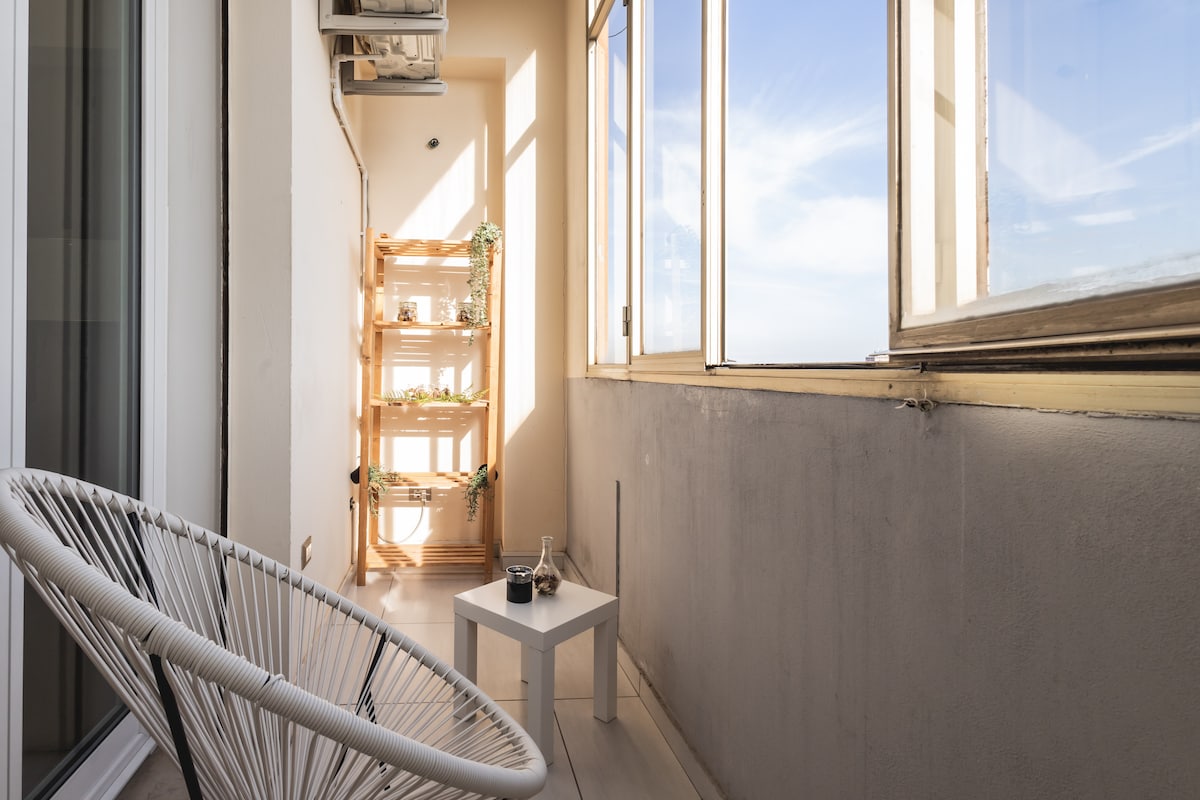 A bright balcony space is presented with large windows allowing natural light to fill the area. An acapulco chair is positioned beside a small white table. A wooden shelf with potted plants adds a touch of greenery to the inviting atmosphere.