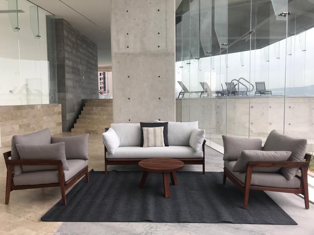 A modern seating area features a light-colored sofa and two armchairs, each adorned with cushions. A round wooden coffee table sits centrally on a textured gray rug. Large glass panels allow natural light to fill the space while providing a view of the outdoor pool area.