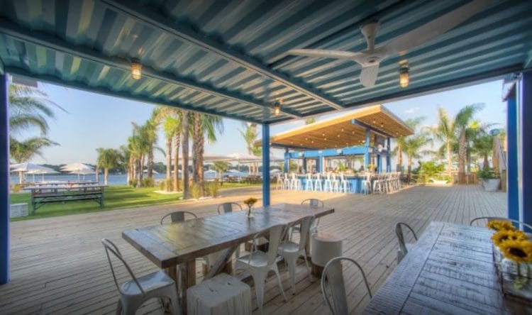 An outdoor dining area features rustic wooden tables surrounded by white chairs under a covered patio. Palm trees and green lawn can be seen, with a pool bar in the background. Soft lighting and a ceiling fan create a relaxed ambiance.