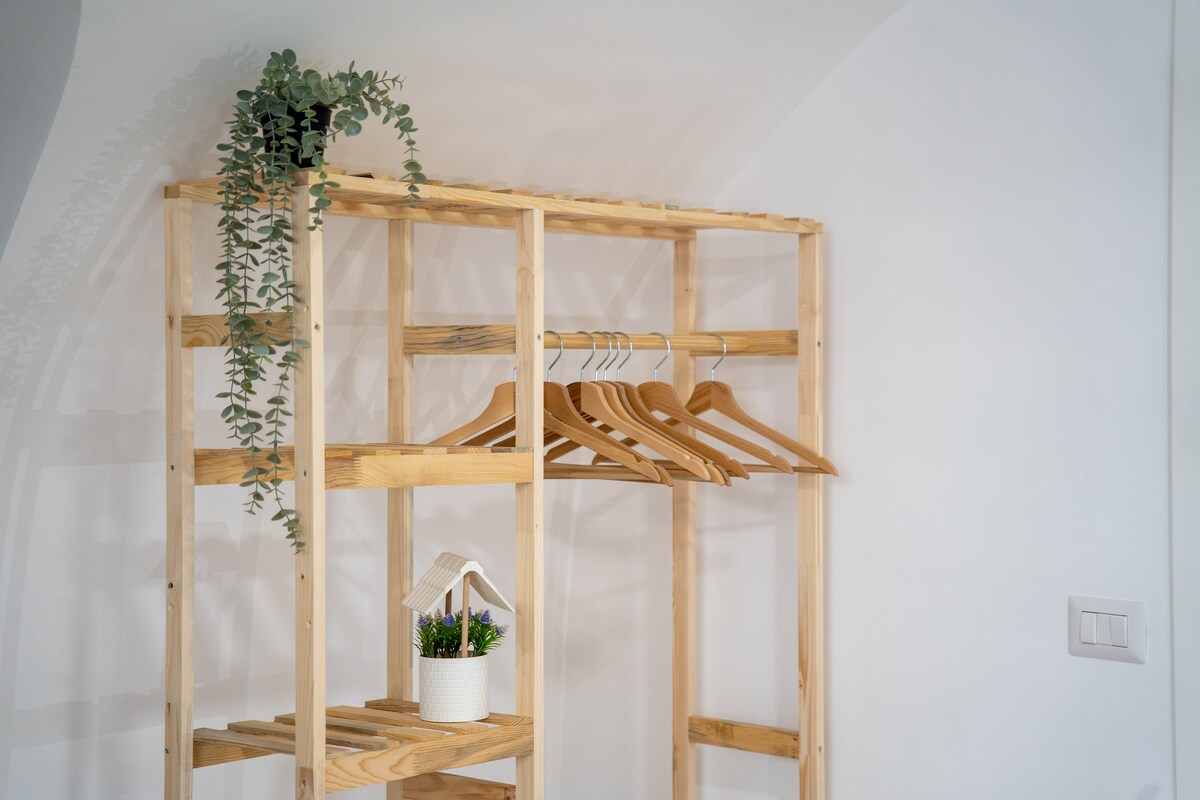 A wooden clothing rack stands against a white wall, featuring several wooden hangers. A small potted plant adds a touch of greenery, while a decorative white planter holds another plant, enhancing the overall aesthetic.