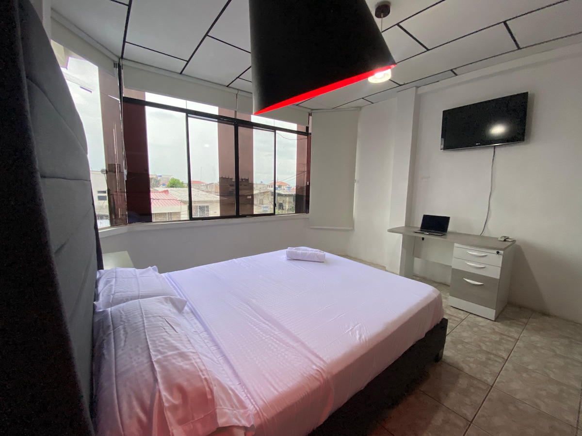 A spacious bedroom features a large, upholstered bed centered against a wall. Natural light filters in through large windows covered by blinds. A flat-screen TV is mounted on the wall, and a desk with a chair is positioned nearby.