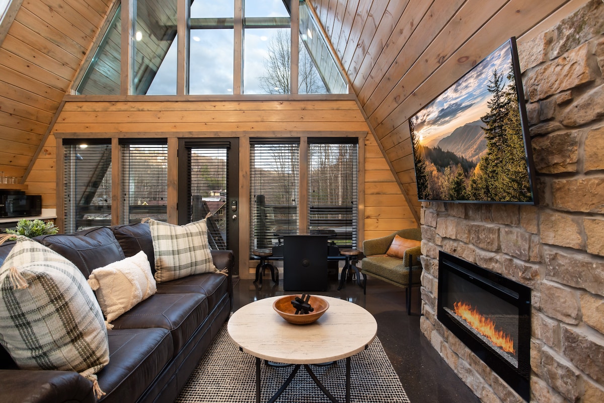 A spacious living area showcases a comfortable leather sofa adorned with patterned throw pillows, complemented by a round coffee table. Natural light filters through large windows, and a mounted TV is positioned above a modern electric fireplace, which is framed by a stone wall.