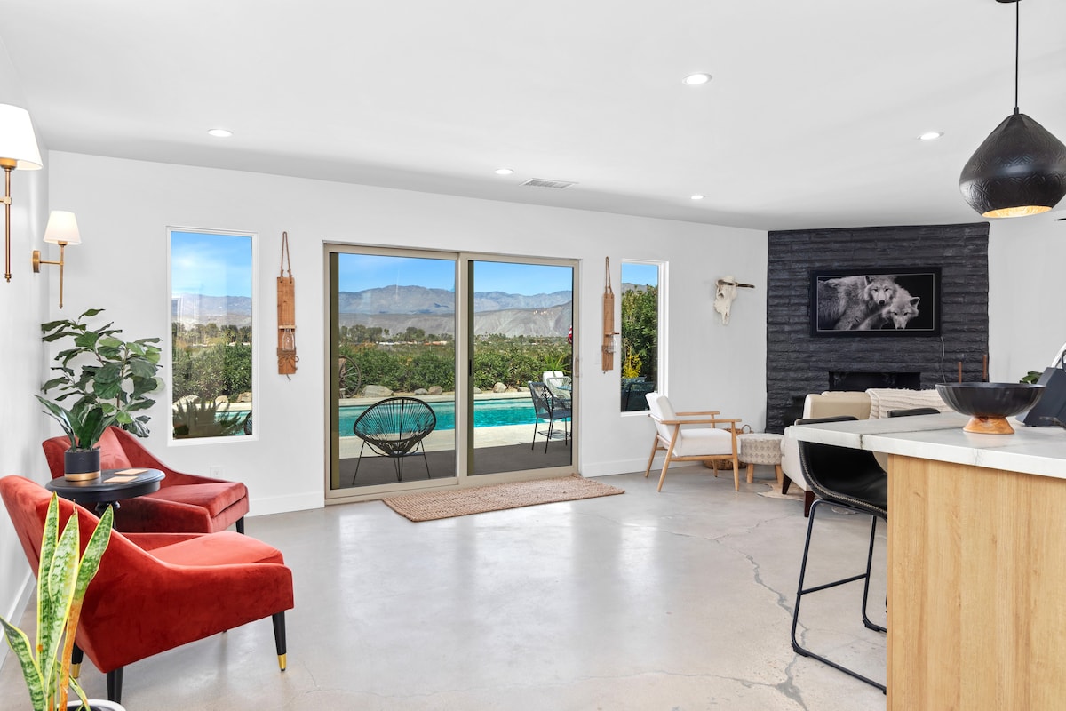A bright living area features large windows that frame views of the pool and surrounding desert landscape. Two red armchairs are positioned on a neutral floor, with a black feature wall enhancing the room's modern aesthetic. A small indoor plant adds a touch of greenery.
