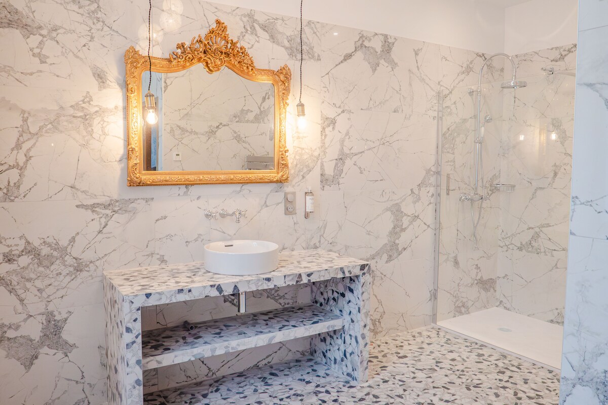 The bathroom features elegant marble walls and floors, complemented by a unique, speckled sink vanity. A large, ornate gold-framed mirror hangs above the sink, while a spacious walk-in shower is equipped with modern fixtures, enhancing the contemporary design.