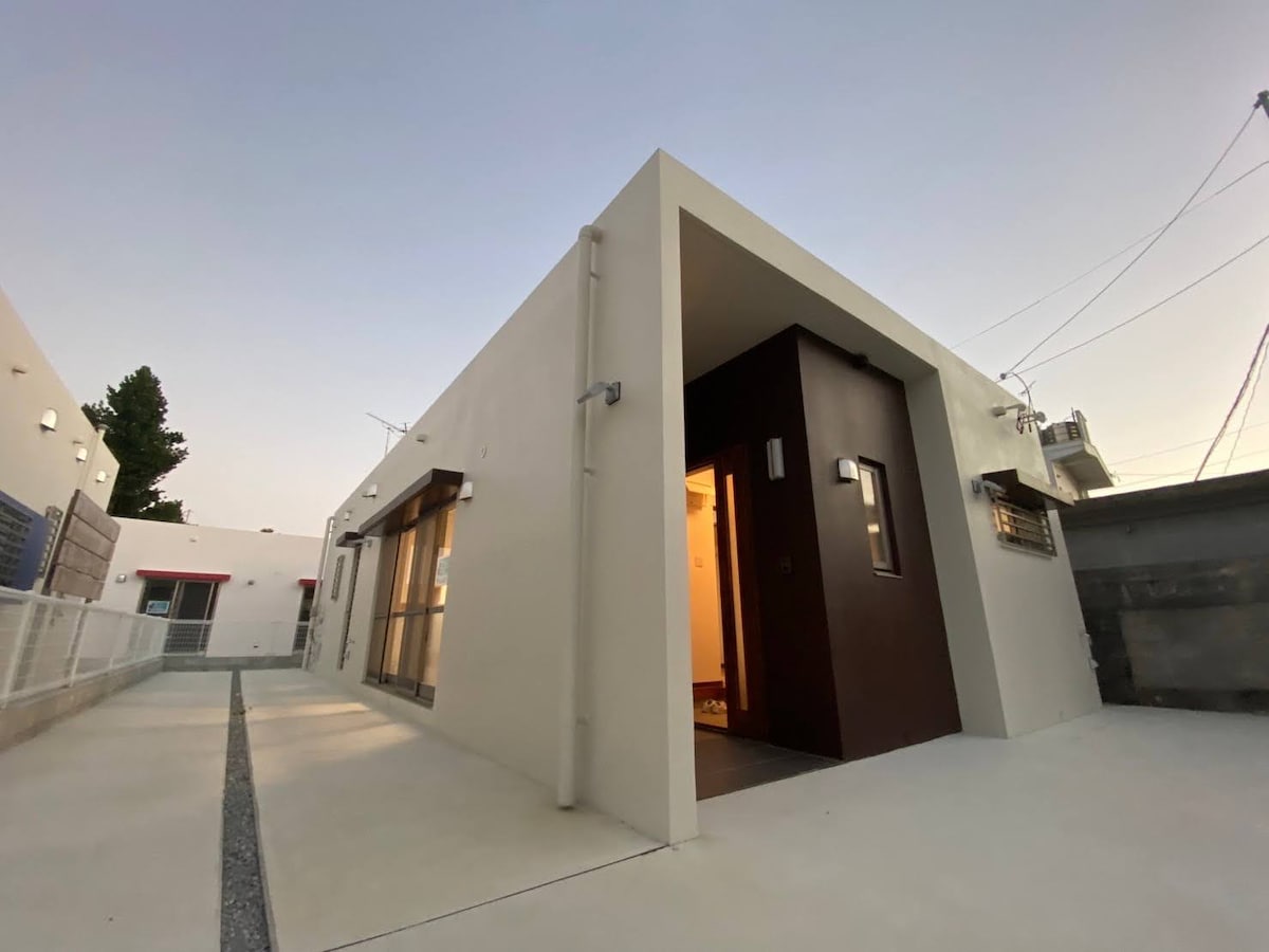 A modern building features a clean, minimalist exterior with a smooth white facade and a dark entrance door. The surrounding area includes a concrete walkway and a landscaped section, enhancing the contemporary design and inviting entrance.