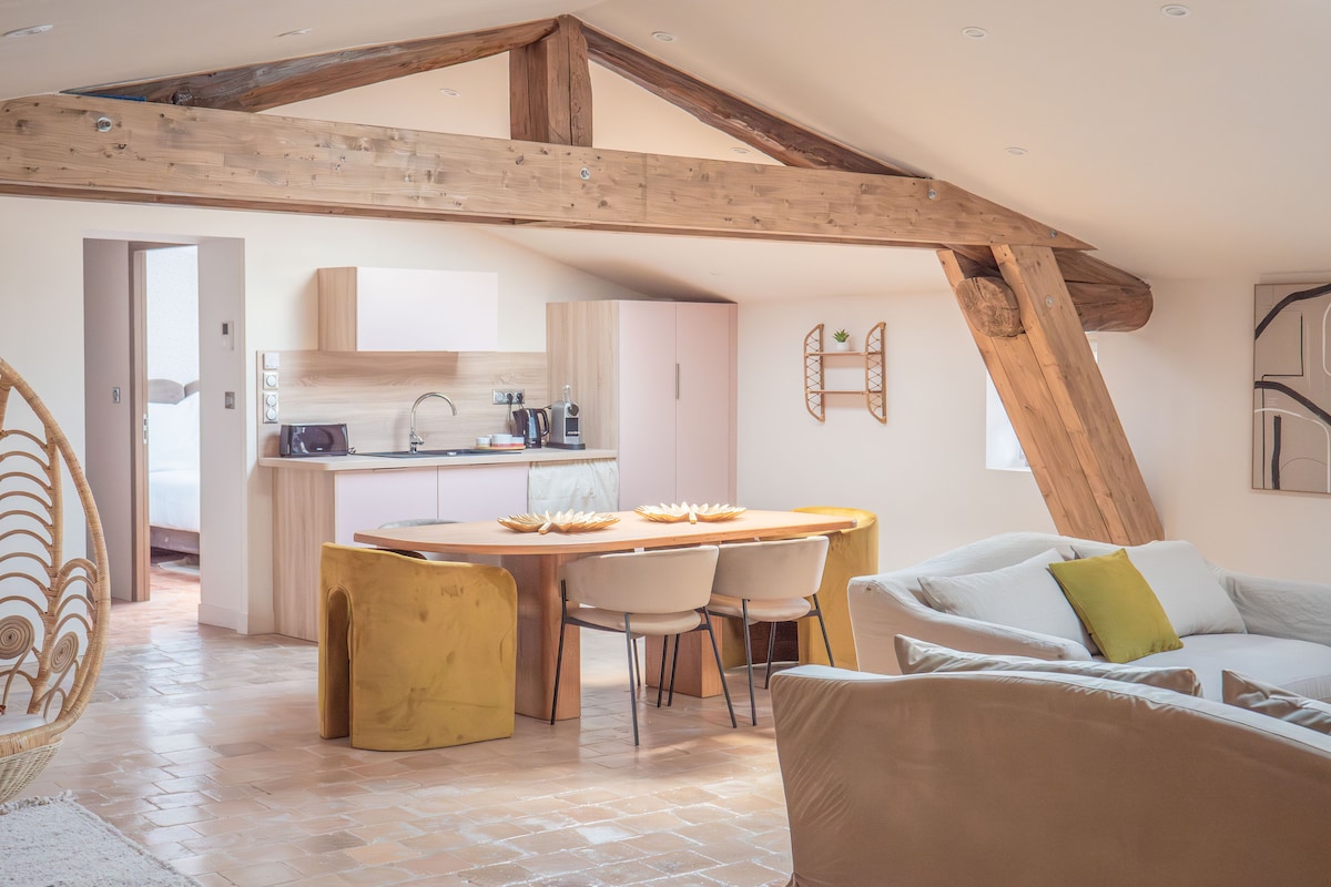 An open living area is illuminated by natural light, featuring wooden beams overhead. A spacious round table with four chairs is centered in the room. A modern kitchen setup is visible in the background, complete with essential appliances and cabinetry.
