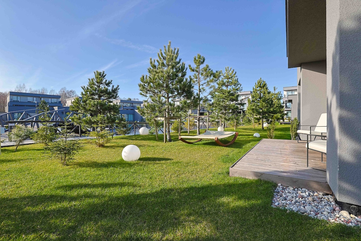 A well-maintained garden is featured, showcasing lush green grass dotted with young trees and decorative white stones. Hammocks are positioned under the trees for relaxation, while modern seating arrangements are visible on a wooden deck adjacent to the living space.