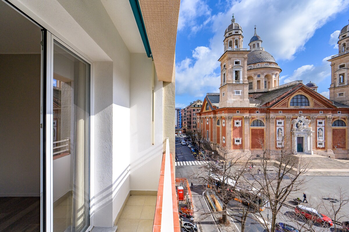 A balcony offers a view of a historic building with prominent architectural details and domed towers. Sunlight casts shadows on the balcony floor, while the street below features trees and vehicles, capturing the vibrant activity of the surrounding area.