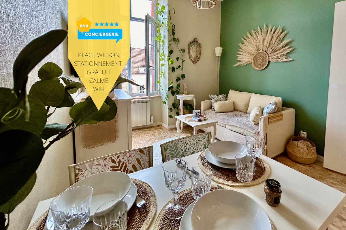 A bright living area features a comfortable light-colored sofa and a decorative wall accent. A dining table is set with elegant tableware, and green plants adorn the space, adding a touch of nature. Large windows allow natural light to fill the room.