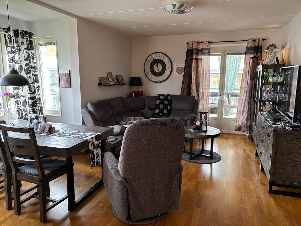 A cozy living area is presented with a plush gray sofa and matching armchair. A round coffee table and a rectangular dining table with wooden chairs occupy the space. Large windows provide natural light, framed by patterned curtains, and personal decorations enhance the surroundings.