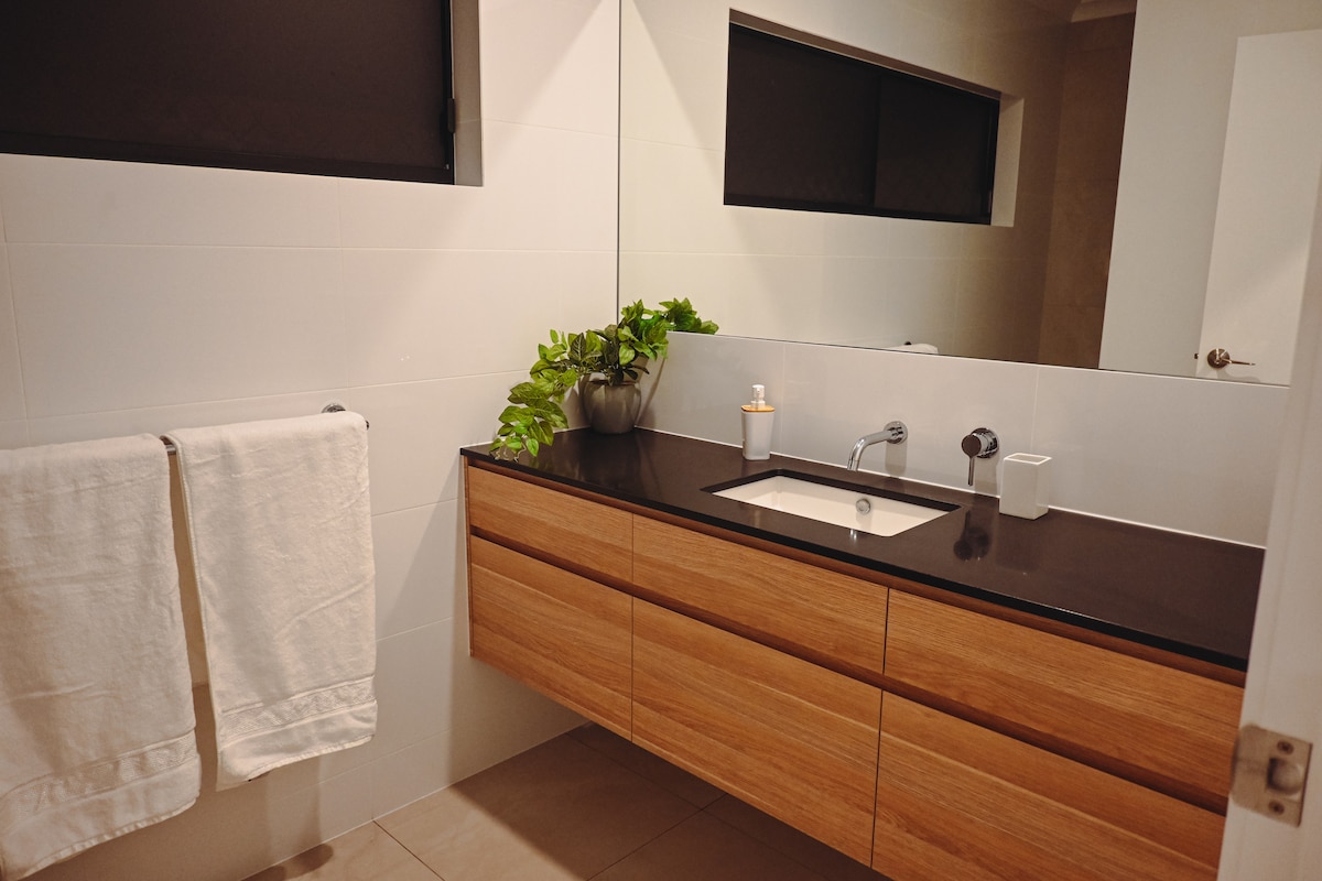 A modern bathroom features a sleek wooden vanity topped with a dark countertop. A rectangular sink is integrated into the surface. Towels are neatly hung on the side, and a potted plant and a candle add subtle decoration. A large mirror reflects the well-lit space.