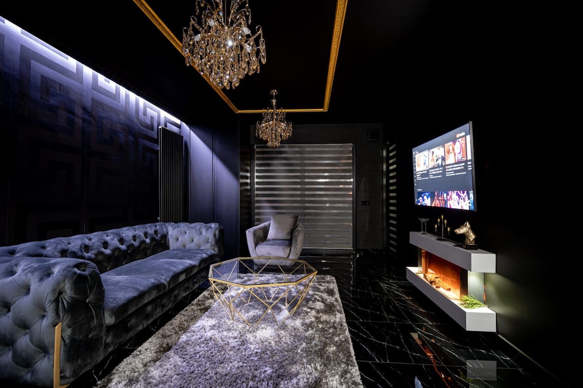 A modern living area features a plush, velvet sectional sofa and a sleek glass coffee table atop a soft area rug. Crystal chandeliers provide ambient lighting against dark walls, while a flat-screen TV is mounted on a stylish feature wall.