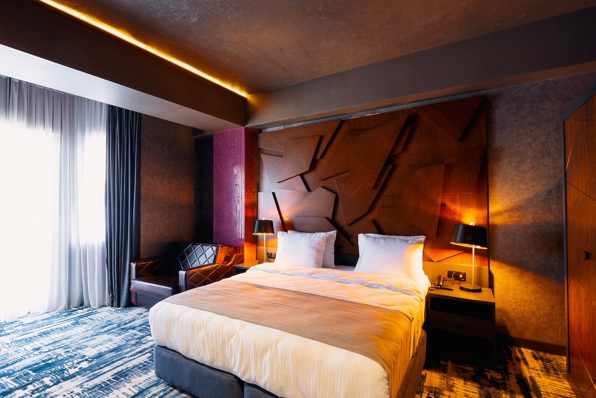 A modern bedroom features a spacious bed with wood paneling behind the headboard. Two bedside lamps provide soft lighting. A comfortable chair sits in the corner, and large windows with sheer curtains allow for natural light.