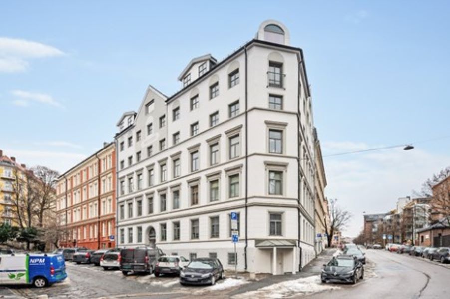A modern building with a white facade is shown on a quiet street. Large windows are positioned throughout the structure, allowing natural light. Surrounding parking is visible, with vehicles lined along the curb. Snow lightly covers the ground, indicating a winter season.