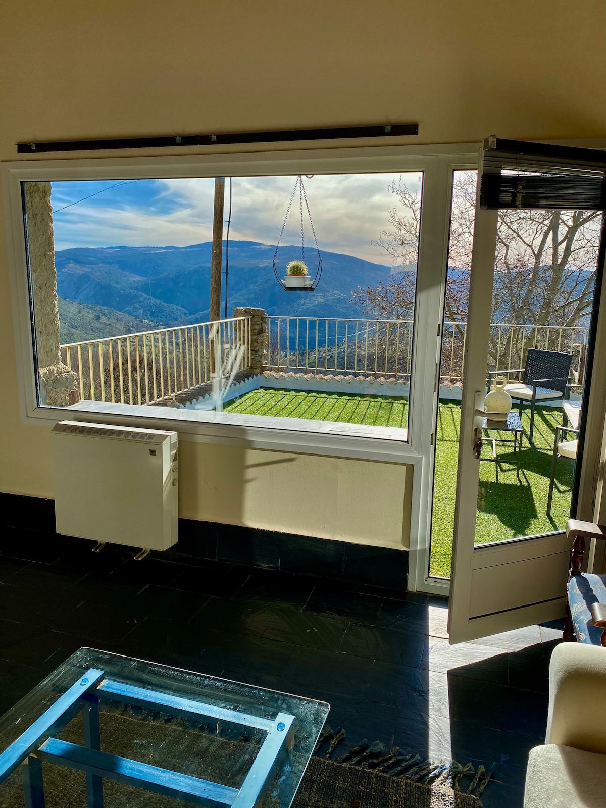 A large window offers breathtaking mountain views, while sunlight fills the open space. A terrace is visible outside, featuring a hanging chair and outdoor seating on green grass. A glass coffee table is positioned within the room, complementing the overall design.