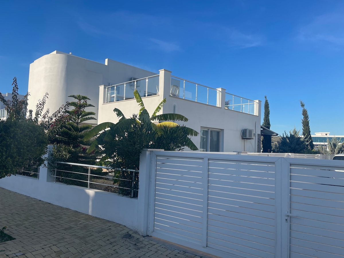 A modern villa exterior is captured against a clear blue sky. Lush greenery surrounds the property, enhancing its serenity. The rooftop terrace with glass railings is visible, providing an inviting outdoor space. A white gated entrance adds to the overall appeal of the villa.