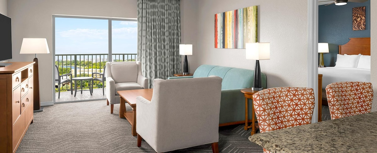An inviting living area showcases a light-colored sofa and comfortable armchairs. A wooden coffee table sits in front, complemented by a textured area rug. Large sliding doors lead to a balcony with views, while colorful artwork decorates the walls.