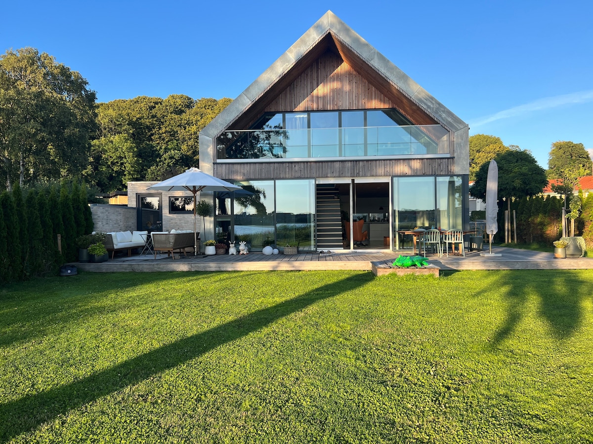 The modern villa features a striking architectural design with a triangular roof and expansive glass windows. A spacious outdoor area includes a lounge set under an umbrella, surrounded by green grass. The tranquility of the surrounding nature and access to the lake are visible.