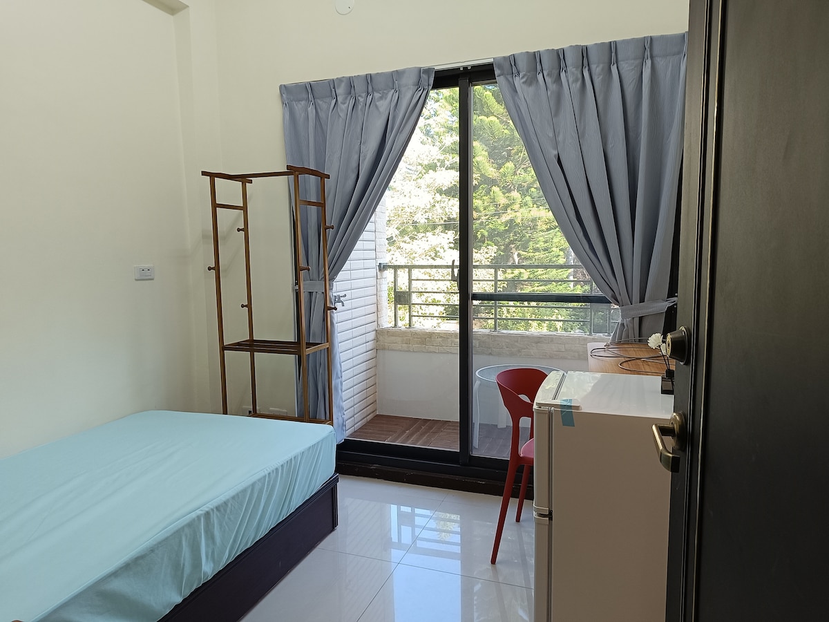 A bright room is showcased featuring a spacious bed with a light blue cover, accompanied by a compact refrigerator. A glass door provides access to a balcony, framed by large curtains. Natural light illuminates the room through the door, highlighting the minimalist decor.