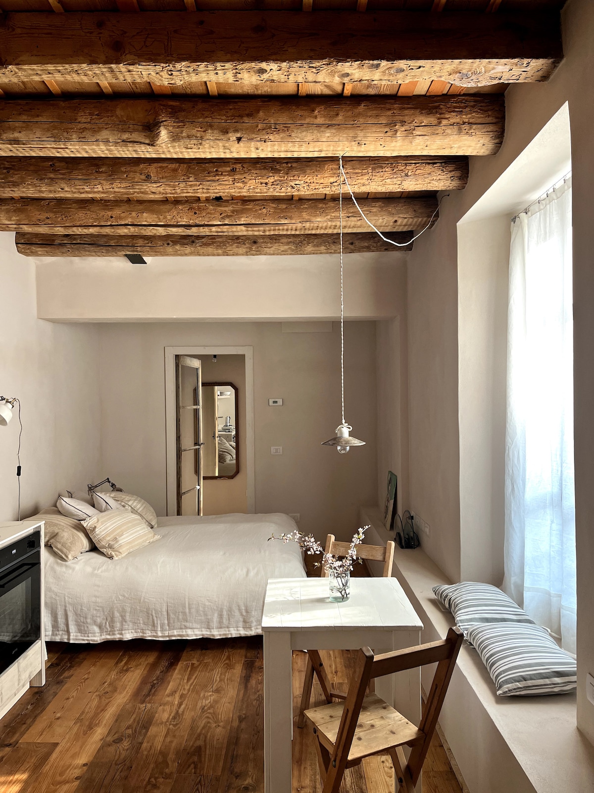 A compact studio features exposed wooden beams that accentuate the high ceiling. A spacious bed is positioned next to a window, while a small dining table with two chairs occupies the area. Natural light filters through sheer curtains, illuminating the neutral tones of the decor.
