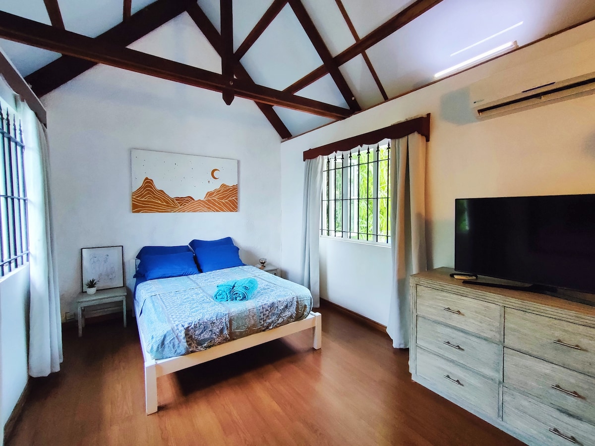 A spacious bedroom features a white bed with blue linens. The wooden ceiling showcases exposed beams, while large windows with dark grills allow natural light to enter. A dresser and a television are positioned against the wall, complementing the room's functional layout.