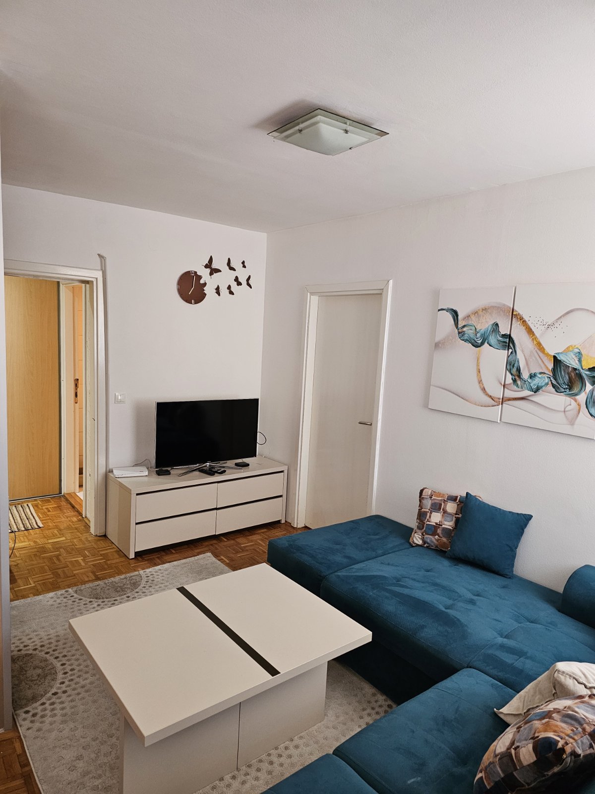 A living area is presented with a blue L-shaped sofa and a light-colored coffee table. A wall-mounted television is visible across from the seating area. Artwork featuring abstract designs decorates the wall, and a patterned rug adds texture to the wooden floor.