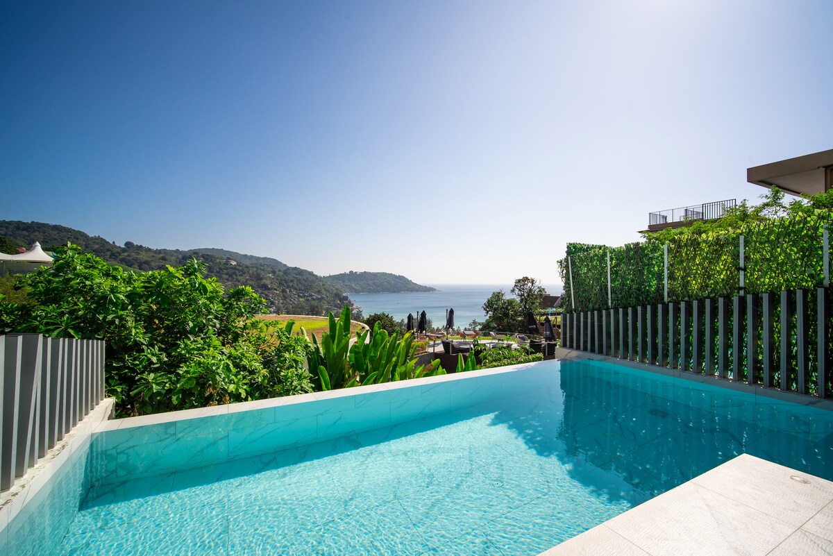 An infinity pool overlooks the scenic coastline, framed by lush greenery. The clear blue waters reflect the vibrant sky, providing a serene setting for relaxation. The horizon showcases a blend of ocean and hills, enhancing the tranquil ambience of the surroundings.