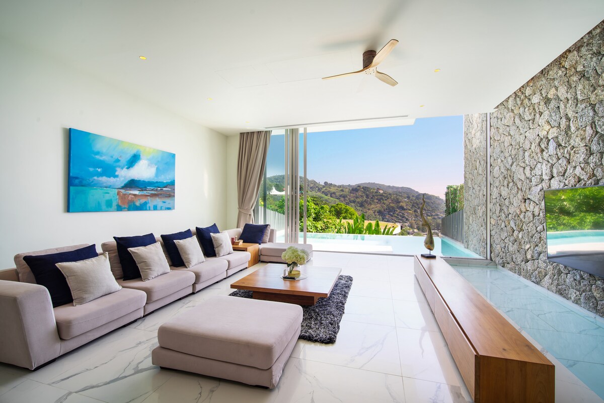 A modern living area is presented with a low large sofa adorned with decorative cushions. Natural light floods the room through expansive glass doors, revealing scenic views of the outdoors. A wooden coffee table centers the space, complemented by minimalist decor and a statement wall.