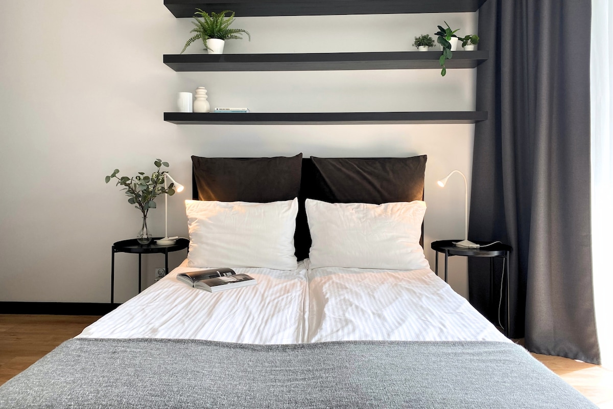 A sleeping area is highlighted by a spacious bed with neutral bedding. Two nightstands are located on either side, each featuring a lamp and a small decorative plant. A minimalist shelving unit above displays various decor items against a light-colored wall.
