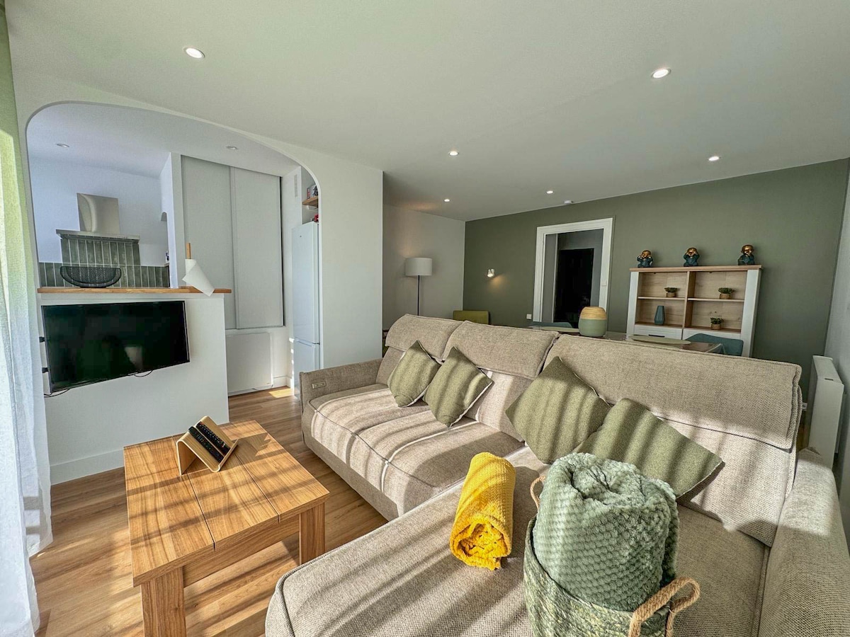 A spacious living area is showcased, featuring a comfortable sectional sofa adorned with green and yellow throw blankets. A wooden coffee table sits in front, and a flat-screen television is mounted on the wall. Soft natural light enters through large windows, creating a bright atmosphere.