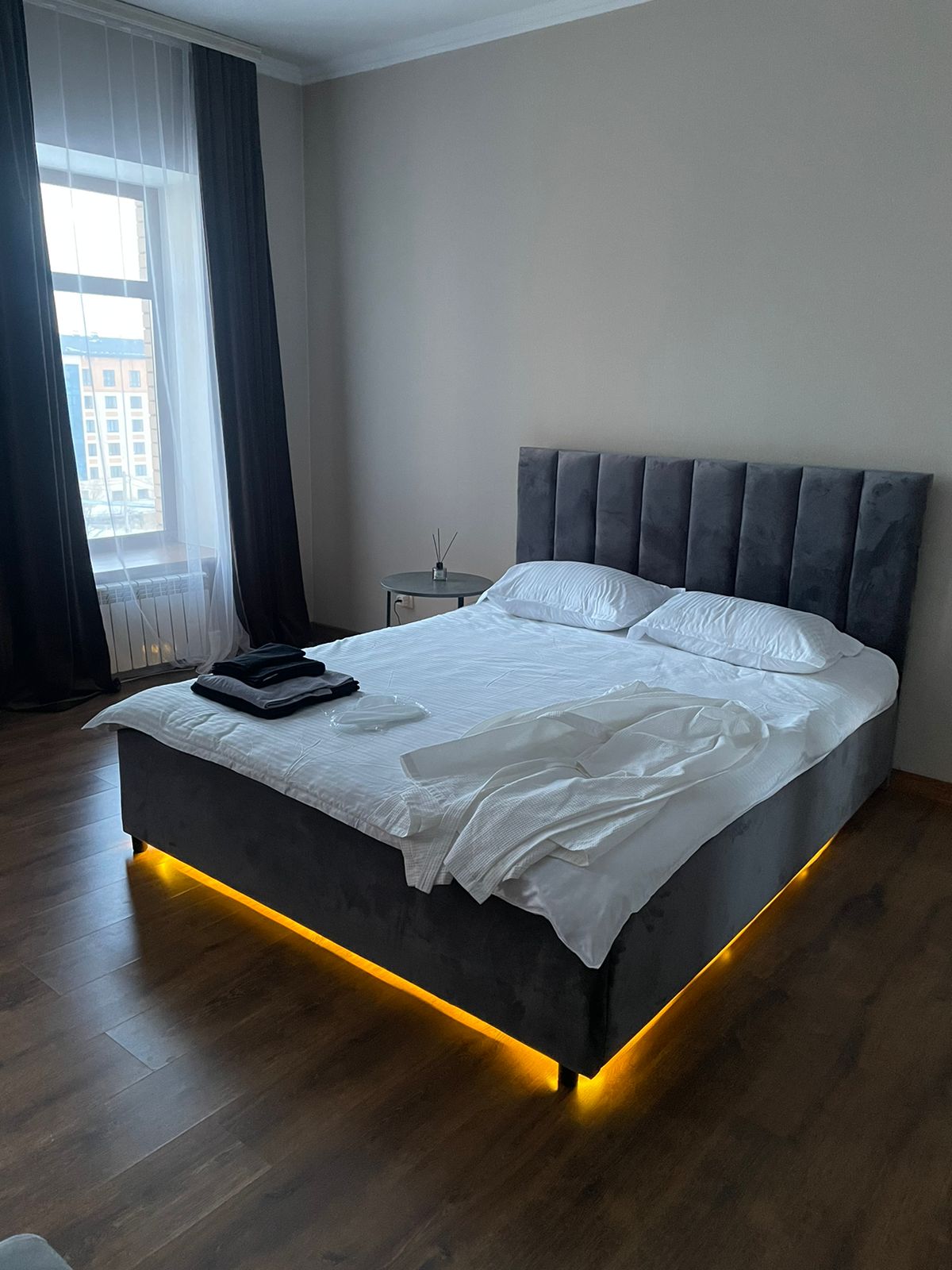 A spacious bed with soft, white linens is centered in the room, complemented by a sleek, grey upholstered headboard. Soft lighting emanates from beneath the bed, creating a warm glow. Natural light enters through a large window, partially covered by dark curtains.