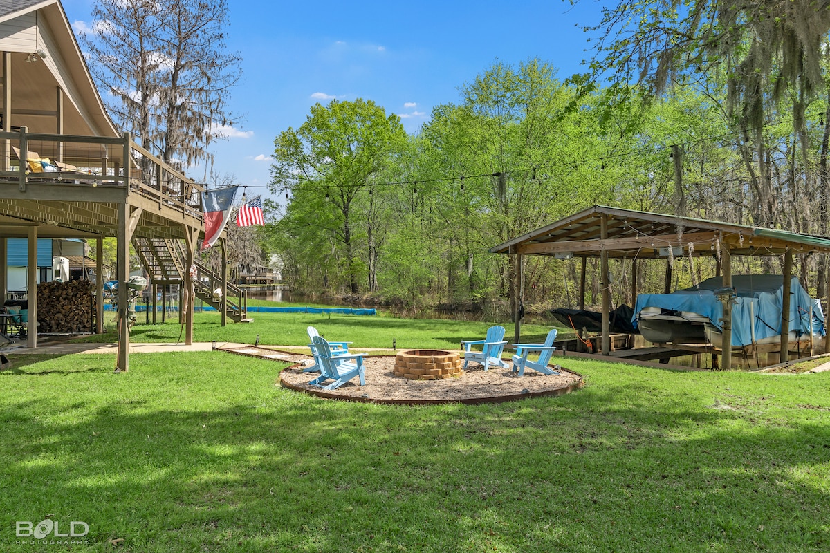 An outdoor area is showcased with a circular fire pit surrounded by blue adirondack chairs, set on a grassy lawn. A covered dock is visible nearby, along with a wooden structure. The backdrop consists of lush trees, creating a serene environment.