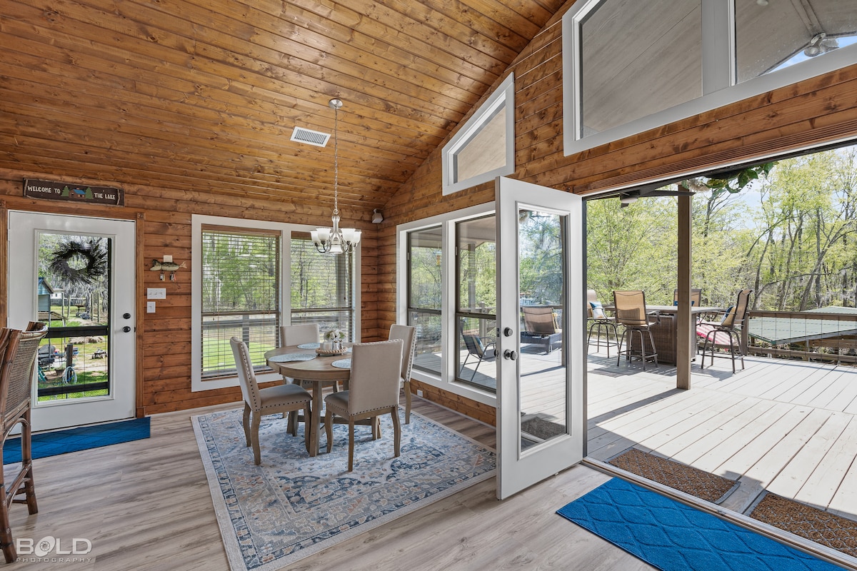 A bright dining area features a wooden table surrounded by upholstered chairs, illuminated by natural light from large glass doors. The room is adorned with a decorative rug, and outdoor seating is visible through the open door, enhancing the connection to nature.