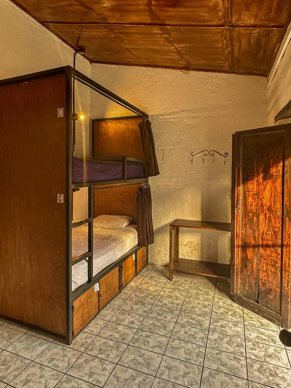 A cozy room is furnished with a wooden bunk bed, featuring a top and bottom sleeping area. Warm lighting is provided by a ceiling fixture. A small wooden table sits against the wall, accompanied by a wardrobe. The tiled floor adds to the room's practical design.