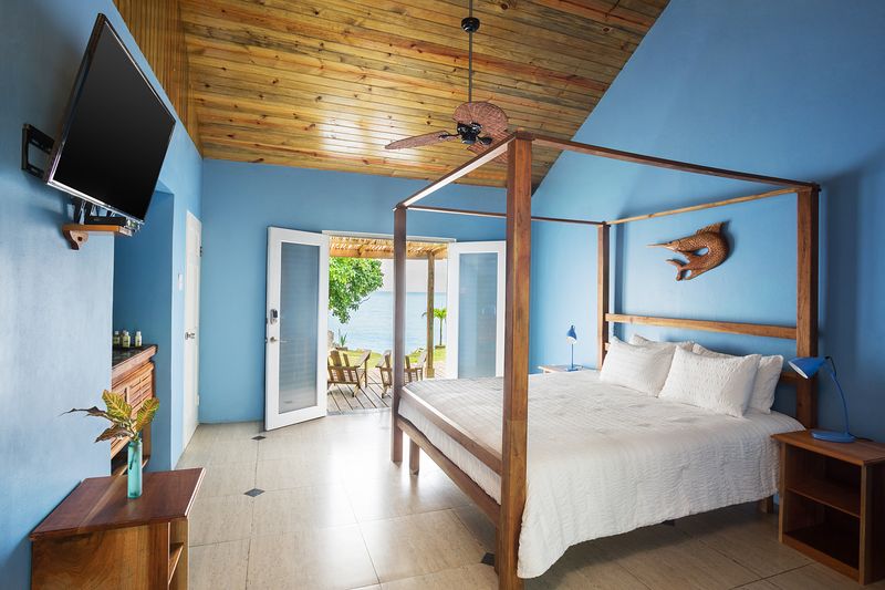 A spacious bedroom features a wooden four-poster bed dressed in crisp linens. French doors open to an outdoor seating area surrounded by greenery. Light wood accents adorn the ceiling, and a flat-screen TV is mounted on the wall opposite the bed.