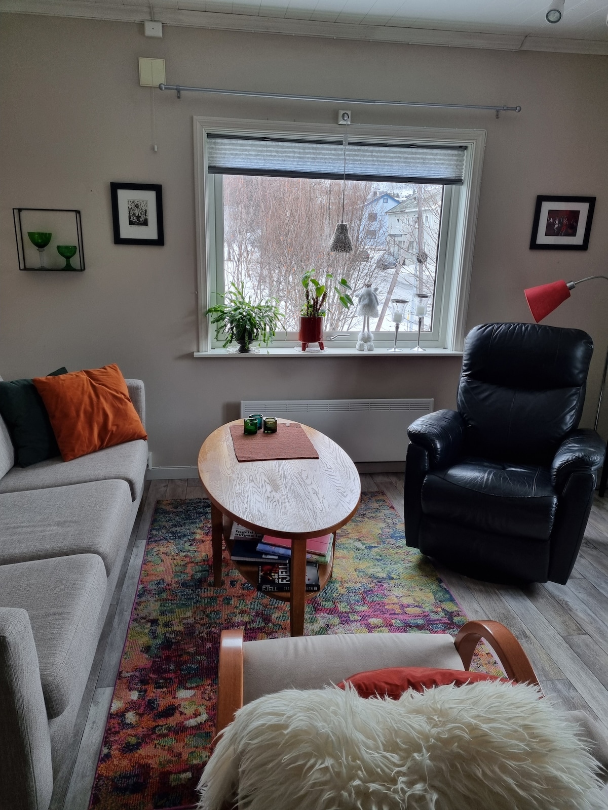 A cozy living room features a light-colored sofa adorned with orange cushions. A circular coffee table is positioned in front of a comfortable recliner. Green plants thrive on the windowsill, framed by sheer curtains. A colorful rug adds warmth to the space.