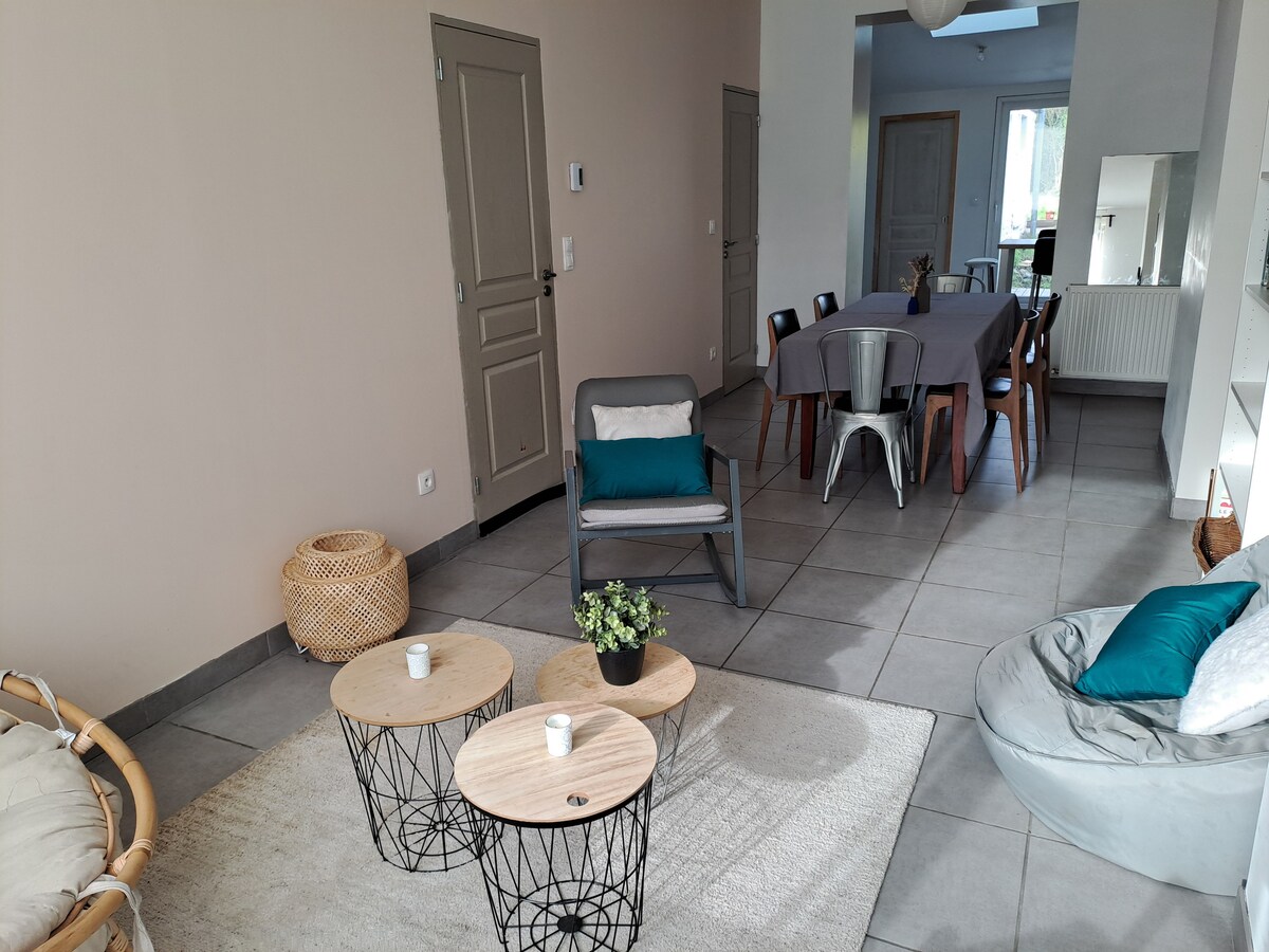 A spacious living area features light-colored walls and gray floor tiles. Two wooden coffee tables are positioned on a soft rug, accompanied by a bean bag chair and a single armchair. A dining table with multiple chairs occupies one side, adding to the multifunctional space.