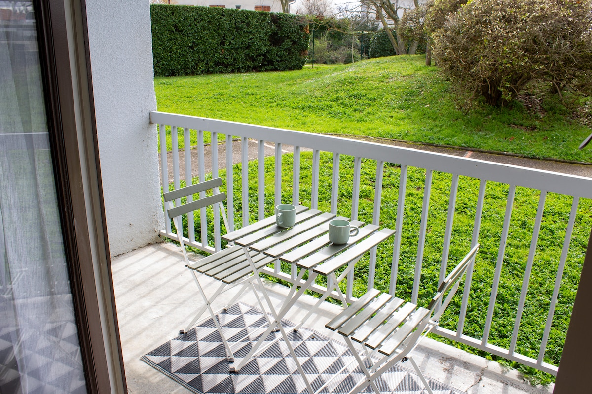 A small terrace is shown, featuring a white folding table with two matching chairs. Two green mugs rest on the table, set against a backdrop of lush grass and greenery visible beyond the railing.