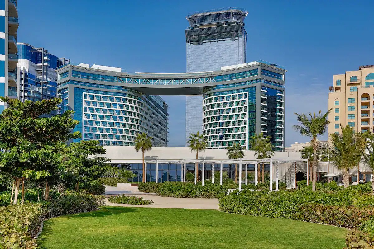 The exterior of the building features modern architecture, showcasing a unique archway connecting two structures. Lush green landscaping surrounds the area, complemented by palm trees and open spaces. A clear blue sky enhances the serene atmosphere of this luxurious location.