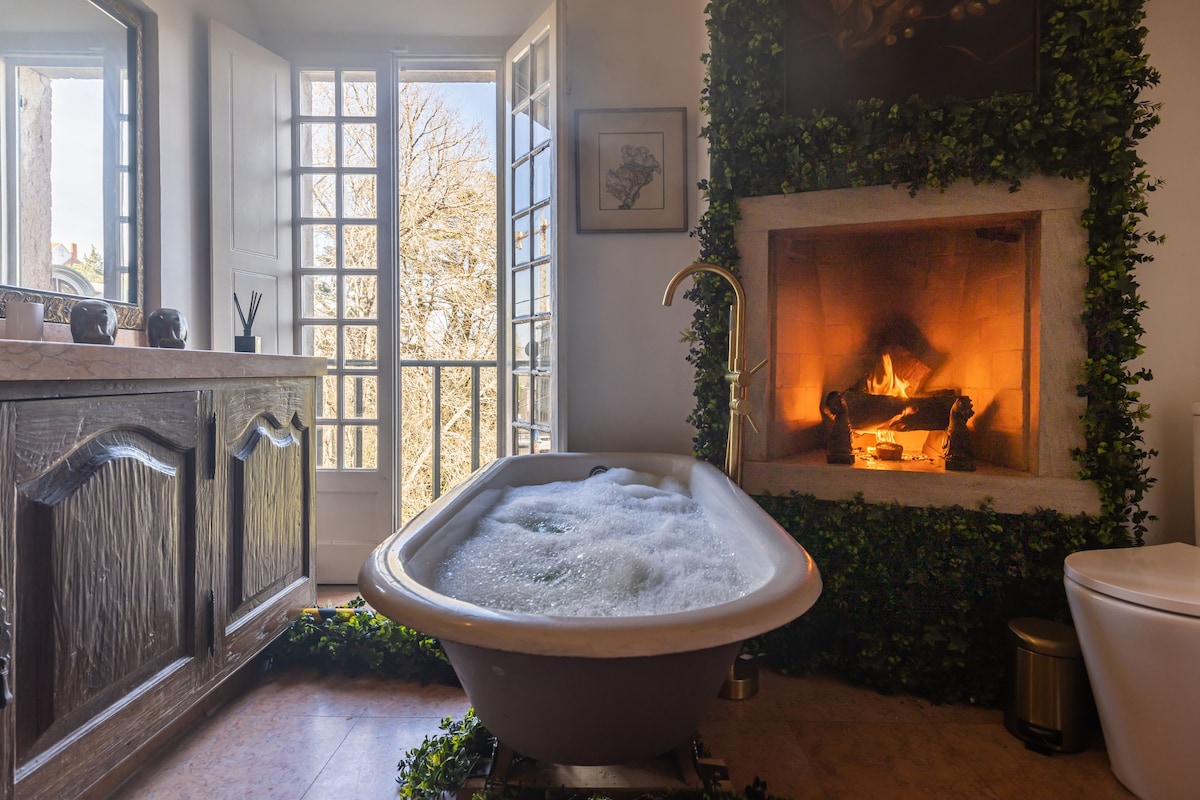 A vintage bathtub filled with bubbles is centrally positioned in a spacious bathroom. A warm fire crackles in a nearby fireplace, framed by green foliage. Large windows allow natural light to illuminate the room, creating a serene bathing environment.