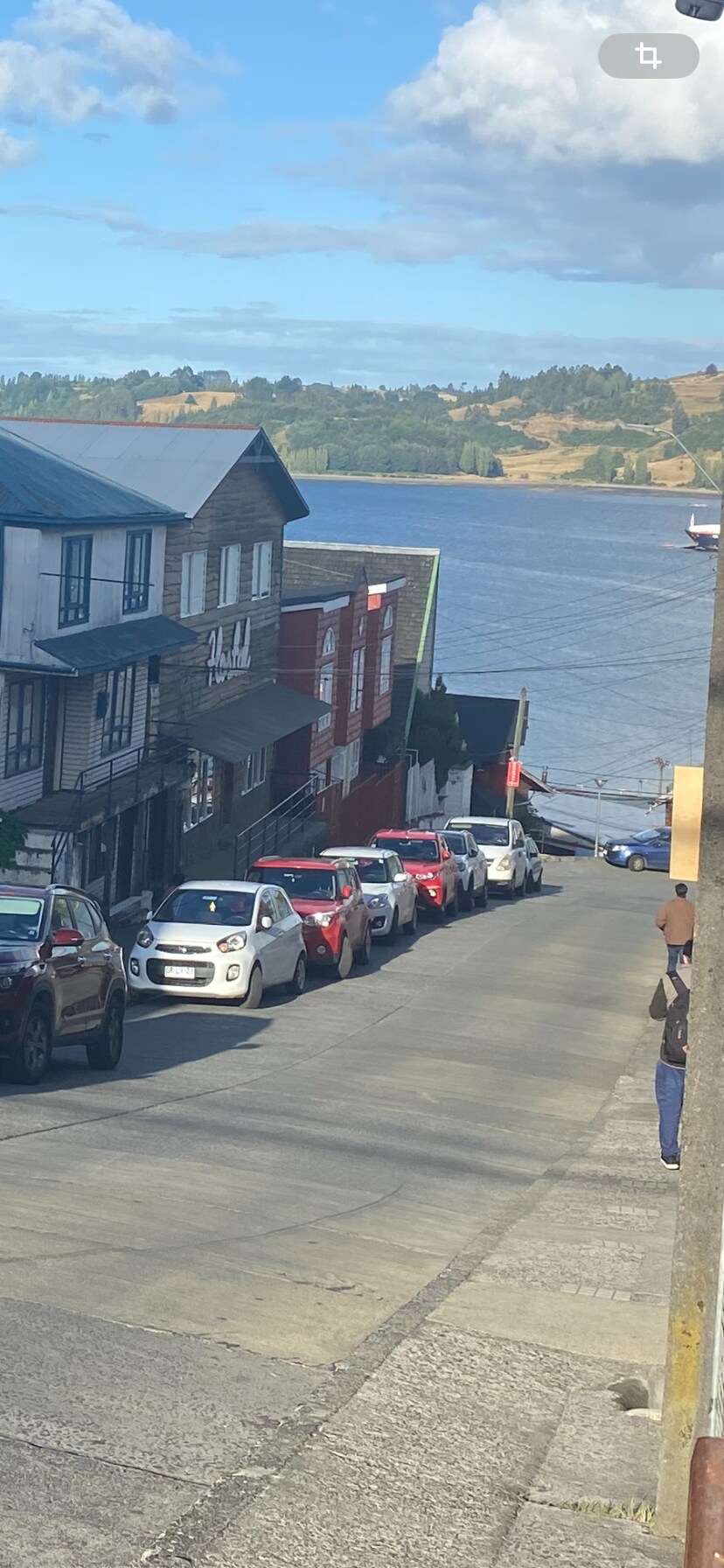 A scenic view of a sloping street lined with various parked vehicles, leading down toward a body of water. The backdrop features a mix of charming buildings with distinct architectural styles, under a partly cloudy sky.