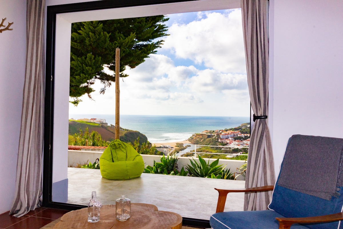 A spacious living area is showcased, featuring large glass doors that open to a stunning view of the ocean and coastline. A comfortable chair is positioned adjacent to a natural wood coffee table. Lush greenery frames the outdoor space, enhancing the overall ambiance.