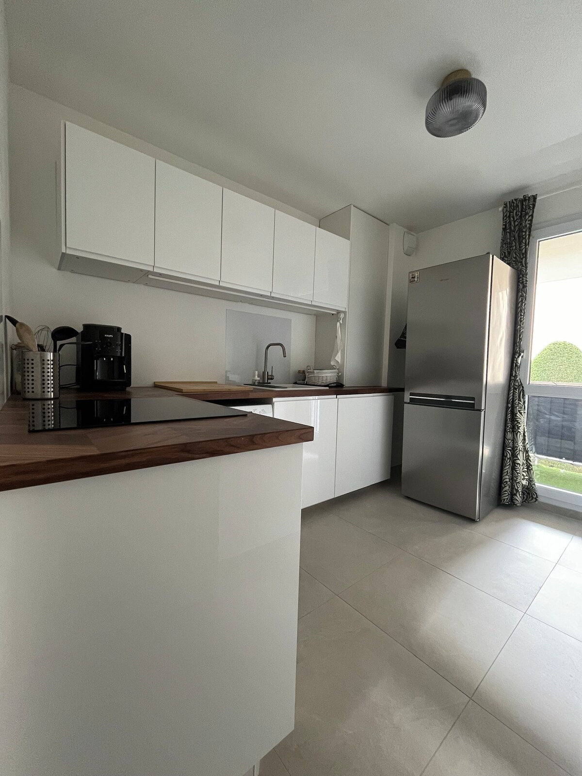 A modern kitchen is presented with white cabinetry and a wooden countertop. Stainless steel appliances, including a refrigerator, are visible, alongside a sink and cooking area. Natural light enters through a glass door that leads to a balcony, enhancing the open feel of the space.