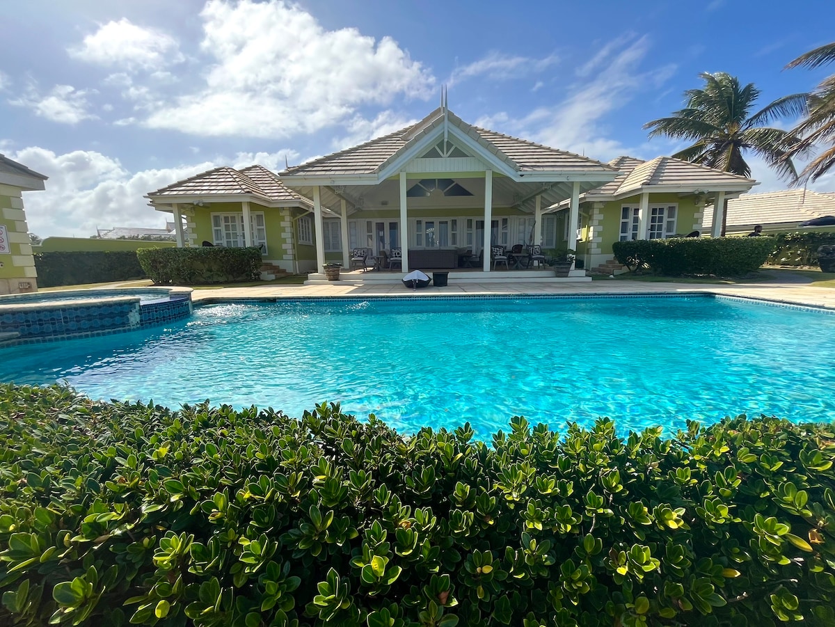 A large villa with pale green accents is viewed from a landscaped area. The expansive swimming pool, featuring clear turquoise water, reflects the sun against a backdrop of palm trees and a partially cloudy sky.