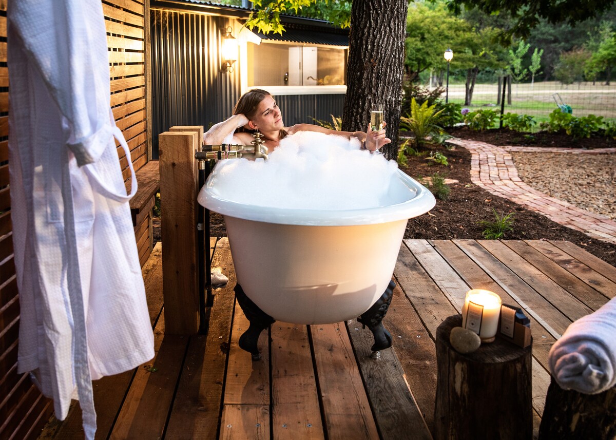 An outdoor claw foot bath sits on a wooden deck, surrounded by lush greenery. Soft, white steam rises from the bath water. A candle rests on a nearby stump, contributing to the serene ambiance. A white robe hangs from a wooden structure in the background.