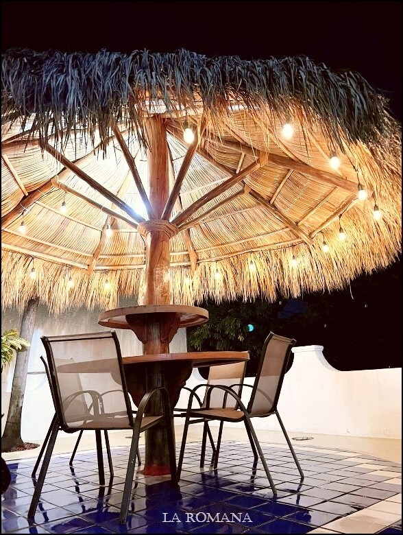 An outdoor palapa with a thatched roof is illuminated by soft lights, creating a cozy atmosphere. A round table is surrounded by four chairs, set against a tiled floor, inviting guests to enjoy meals or gatherings in a tranquil setting.