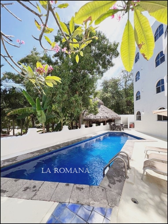 A serene outdoor pool is framed by lush tropical foliage and vibrant flowers, providing a refreshing and inviting space. The pool area is designed with stone paving and is adjacent to a thatched-roof palapa, offering a relaxed setting for guests.