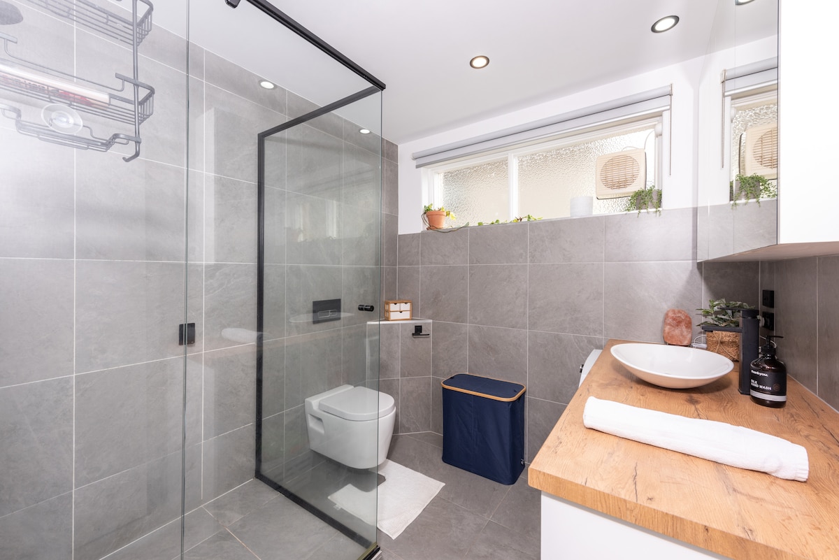 The bathroom features a sleek design with gray tiled walls and flooring. A glass shower enclosure stands adjacent to a modern toilet. A wooden countertop holds a round basin, accompanied by neatly arranged toiletries. Natural light enters through a window adorned with potted plants.
